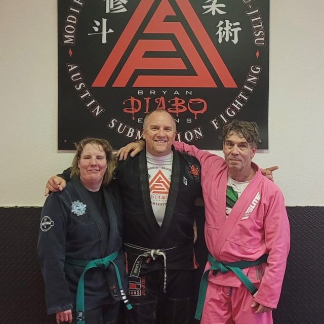 Nikki, Bryan and Ken pose in front of the academy's sign. Each wears a Gi and their belt.