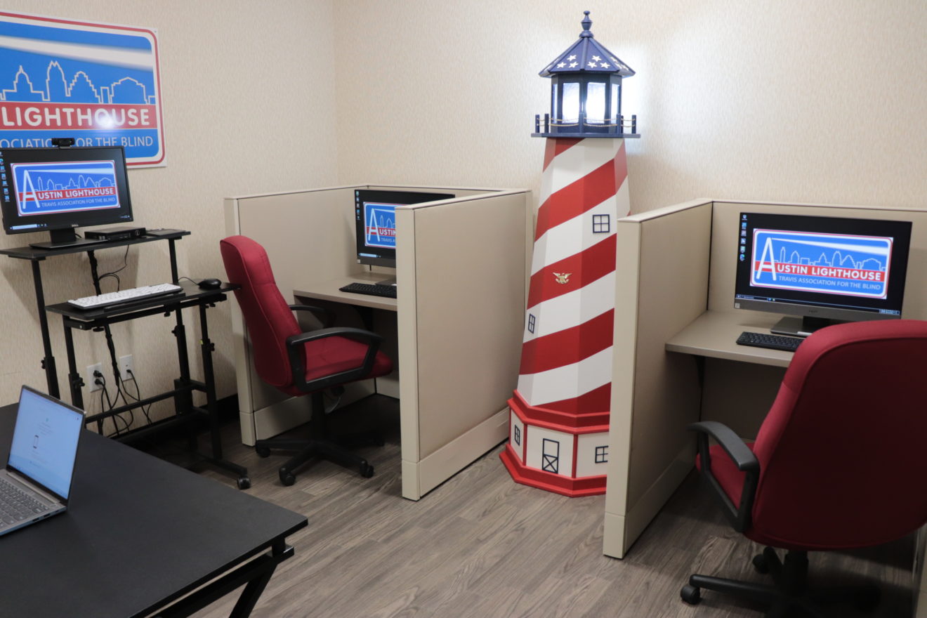 Two desks with computers are pushed against a wall. The lighthouse logo is on the computer screens.