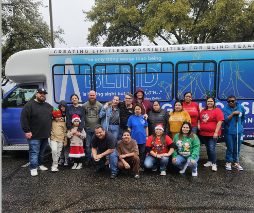 A group of men, women and children pose in front of the Austin Lighthouse bus in a parking lot wet from rainfall. The sky is cloudy.